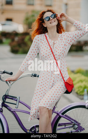 Active cheerful woman rides bicycle in city, wears stylish sunglasses, has toothy charming smile, dressed in summer dress, enjoys sunny day, spends fr