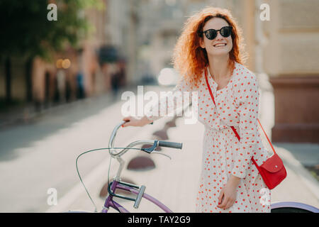 Smiling red haired European woman has glad expression, stands near bicycle, carries red bag, focused somewhere into distance, poses over blurred backg Stock Photo