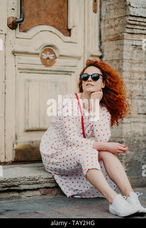 Red haired thoughtful woman has rest after strolling, poses near old building, sits on threshold, wears sunglasses, dress and white sneakers, explores Stock Photo