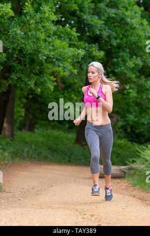 A fit, modern, caucasian woman, running in a park, along a path, wearing a pink crop top, grey leggings and training shoes. Blonde with a pony tail Stock Photo