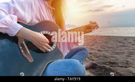 woman's hands playing acoustic guitar, capture chords by finger on sandy beach at sunset time. playing music concept Stock Photo