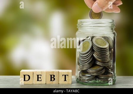 Liabilities and savings for payment concepts. A man hand putting coin into full stacked coins in glass jar on wood table with blurred nature backgroun Stock Photo