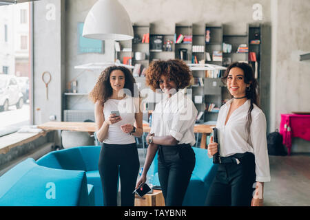 three young multicultural businesswomen standing with smartphone in modern loft – inventive, freelance, dedication Stock Photo