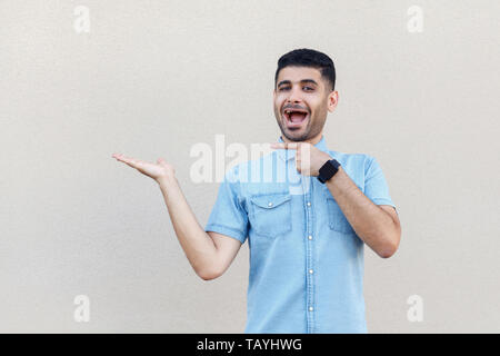 Portrait of surprised handsome young bearded man in blue shirt standing, holding, pointing something and looking at camera with amazed face. indoor st Stock Photo