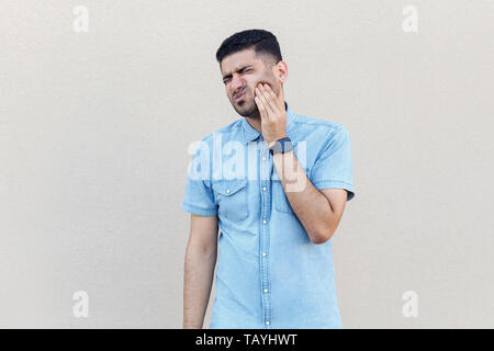 Tooth pain. Portrait of worry sad handsome young bearded man in blue shirt standing, touching his cheek because feeling toothache. indoor studio shot  Stock Photo