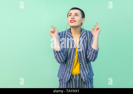 Portrait of hopeful handsome beautiful short hair young stylish woman in casual striped suit standing, crossed fingers and wishful to win . indoor stu Stock Photo