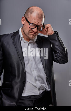 Depression bald business man in eyeglasses scratching the head in suit on grey background. Closeup studio portrait. Stock Photo