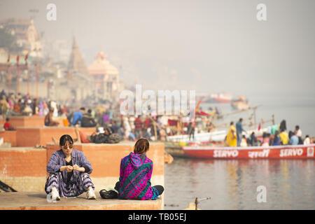 Two Chinese tourists are sitting on a Ghat in Varanasi. Manikarnika Ghat (Burning Ghat) in the background. Varanasi, India. Stock Photo