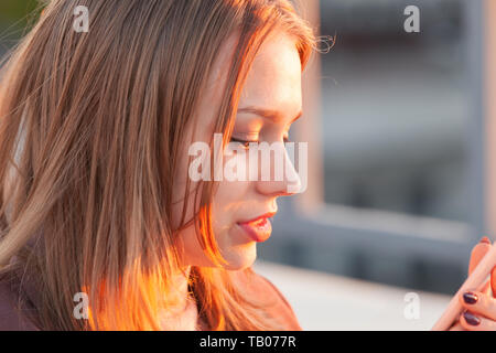 Beautiful blond teenage girl with a smartphone, close up outdoor portrait with back-lit sunlight Stock Photo