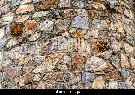 The motley background of the facade of the old oval tower of stones of different breeds and shades of untreated natural stone Stock Photo