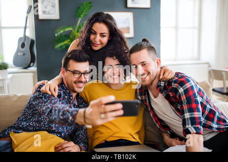 A group of young friends with smartphone sitting on sofa indoors, taking selfie. Stock Photo