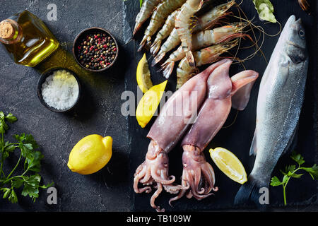 close-up of raw whole sea bass, king prawns, calamari, lemon slices, spices and herbs on a slate plate on a concrete table, view from above Stock Photo