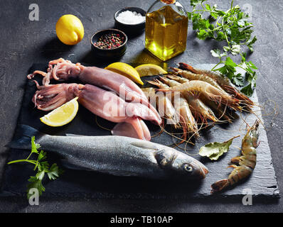 close-up of sea bass, shrimps, calamari, lemon slices, spices and herbs on a slate plate on a concrete table, view from above Stock Photo