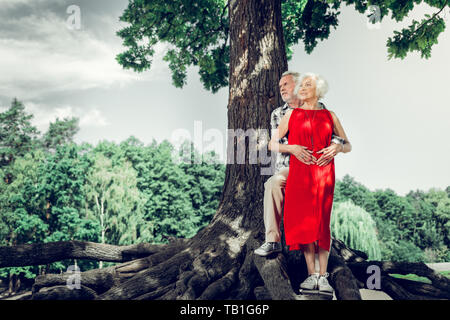 Attractive man tenderly cuddling wife while standing under the tree Stock Photo