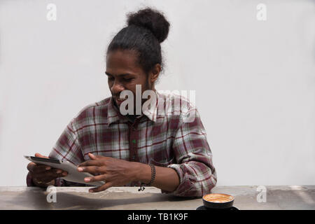 a young black man smiling using tablet and smartphone with glass of coffee, a papua young man using tablet with a coffee cup Stock Photo