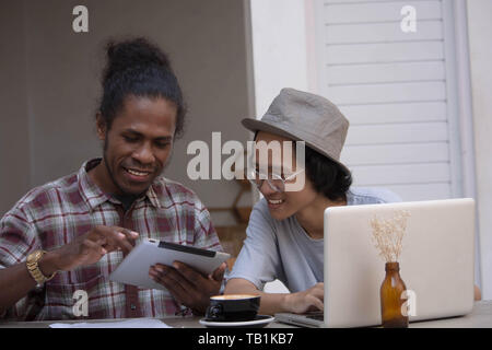 two young creative man discuss with laptop and tablet, young asian and black man working with tablet and laptop in a cafe, papua and asian man working Stock Photo