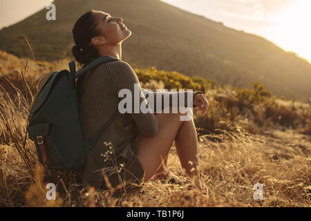 Young woman hiking in countryside sitting and taking rest. Female hiker resting on dry grass field.