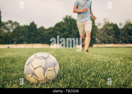 Soccer ball on the grass on a sports field with an anonymous young man in the background running towards it in a low angle view Stock Photo