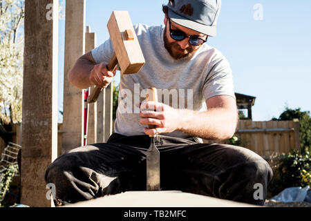 Man wearing baseball cap and sunglasses on building site, using mallet and chisel, working on wooden beam. Stock Photo