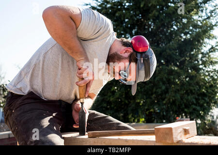 Man wearing baseball cap, sunglasses and ear protectors on building site, working on wooden beam. Stock Photo
