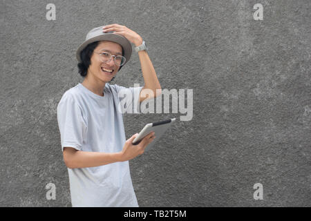 young atractive creative asian man with fedora hat and glasses using tablet and smiling Stock Photo
