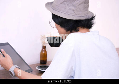 Back view. Young creative man in white shirt and padora hat sitting at table, using laptop while holding a cup of coffee Stock Photo