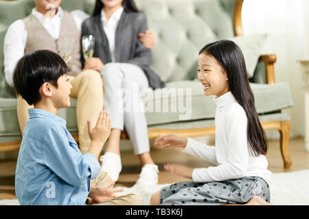 two cute little asian kids brother and sister sitting on carpet playing game with parent sitting on sofa in the background Stock Photo