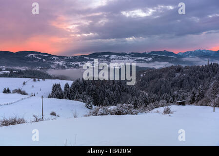 Majestic sunset over a beautiful snowy mountain scenery with a forest partly hidden by fog, meadows and a mountain village Stock Photo