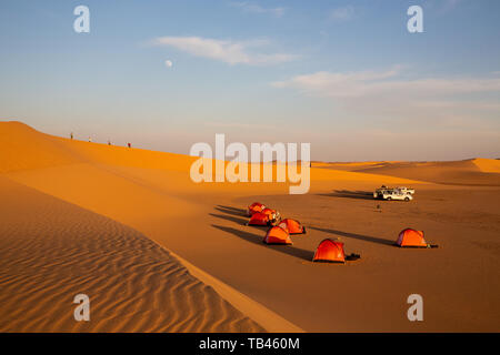 tents pitched for wild camping in the evening sunshine in the western desert of Sudan. Four figures climb the ridge of a sand dune as the moon sets Stock Photo