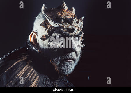 Demon with golden collar on black background. Man with thorns or warts, face covered with glitters. Senior man with white beard dressed like monster Stock Photo