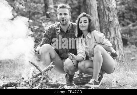 Couple roasting sausages on sticks nature background. Couple in love enjoy camping forest roasting sausage at bonfire. Camping and picnic. Traditional roasted food as attribute of picnic. Stock Photo