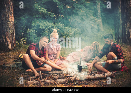 Two couples having picnic in woods. Bearded man and his best friend cooking sausages over fire. Young hikers around campfire in the evening Stock Photo