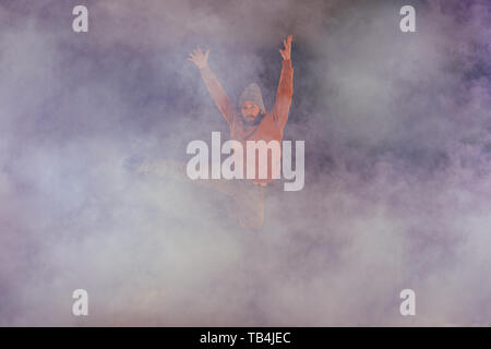 Male breakdancer jumps off high in the air while performing a cool dance move, a lot of smoke in the background. Stock Photo