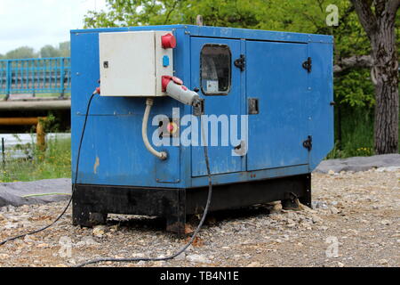 Heavy enclosed industrial power generator located next to river providing electrical power to large water pumps pumping flood water Stock Photo