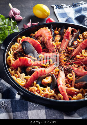 Spanish Fideua, a noodle Paella with seafood - king prawns, white fish meat, calamari, mussels in a black pan on a concrete table with ingredients, ve Stock Photo