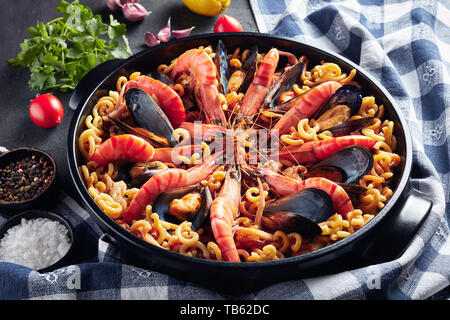 Spanish Fideua, a noodle Paella with seafood - king prawns, white fish meat, calamari, mussels in a black pan on a concrete table with ingredients, ho Stock Photo
