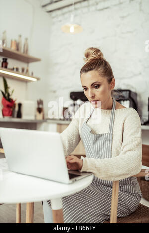 Deep in work. Concentrated smart workwoman of a small cafe sitting in the kitchen working on the laptop. Stock Photo