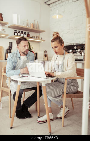 Working side by side. Two focused business partners sitting in front of kitchen equipment in their cafe and working. Stock Photo