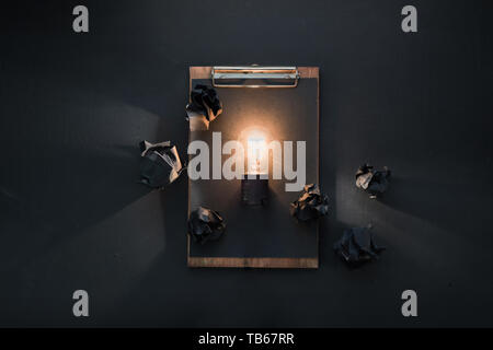 Light bulb shining in black flat lay with crumpled paper balls and a clipboard. The idea for engineering, building or construction concept with copy s Stock Photo