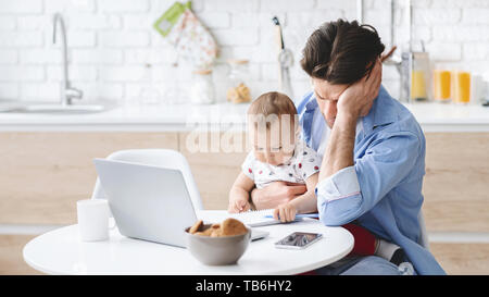 Millennial man tired of working and taking care of baby, covering face in despair. Little boy playing with notebook, kitchen interior, free space Stock Photo