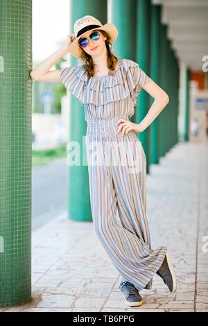 Below view of fashionable teenage girl dressed in green jumpsuit wears  white shoes has trendy hairstyle poses against modern building looks  confident at camera. Young people and fashion concept Stock Photo |