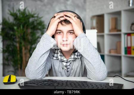 Oops, failure, loss. Teenager boy lost in a computer game. A schoolboy sits upset in front of a computer monitor, covering his face with his hands. Stock Photo