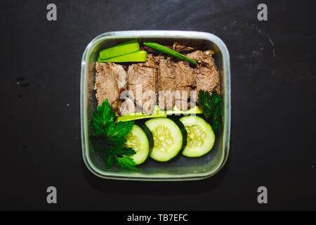 Boiled beef and pieces of fresh cucumber in a small container on a black background, top view, close up. Healthy food, diet, fitness. Stock Photo