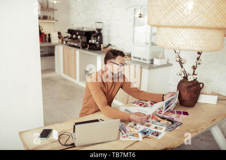 Analysing work. Serious fashion designer sitting at the table looking at his sketches and pictures of dresses comparing them. Stock Photo