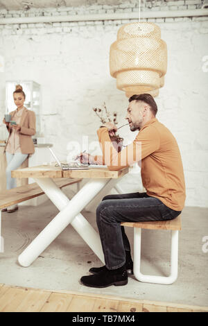 Enwrapped in thoughts. Concentrated fashion designer sitting at the table with eyeglasses in his mouth and thinking about his new clothes. Stock Photo