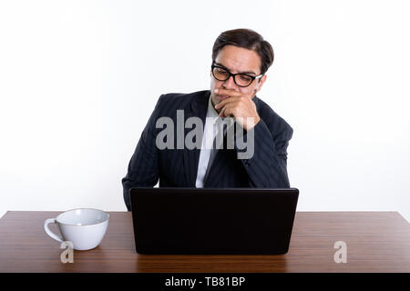 Studio shot of young Persian businessman wearing eyeglasses while thinking and using laptop with coffee cup on wooden table against white background Stock Photo