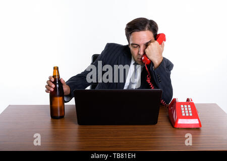 Studio shot of young Persian businessman looking drunk while holding bottle of beer and old telephone with laptop on wooden table against white backgr Stock Photo