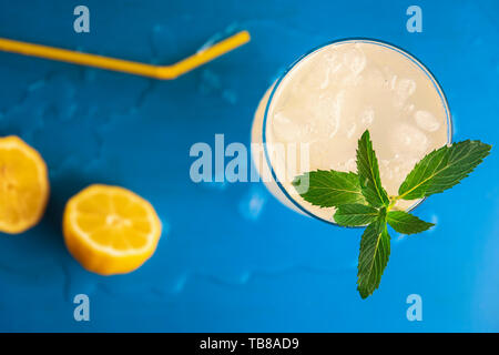 Above view of a lemonade glass with crushed ice and peppermint branch and yellow straw and lemons, on a blue background. Refreshing cold summer drink. Stock Photo