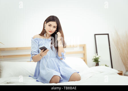 A beautiful Asian woman with headphone relaxing at home , sitting on the bed listening to music from application on mobile phone Stock Photo