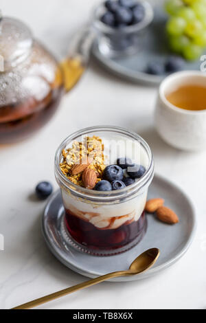 Close up cereal with blueberries and almond, jam and yogurt in glass jar at gray plate on white marble background. Honey, tea, spoon and napkin with b Stock Photo